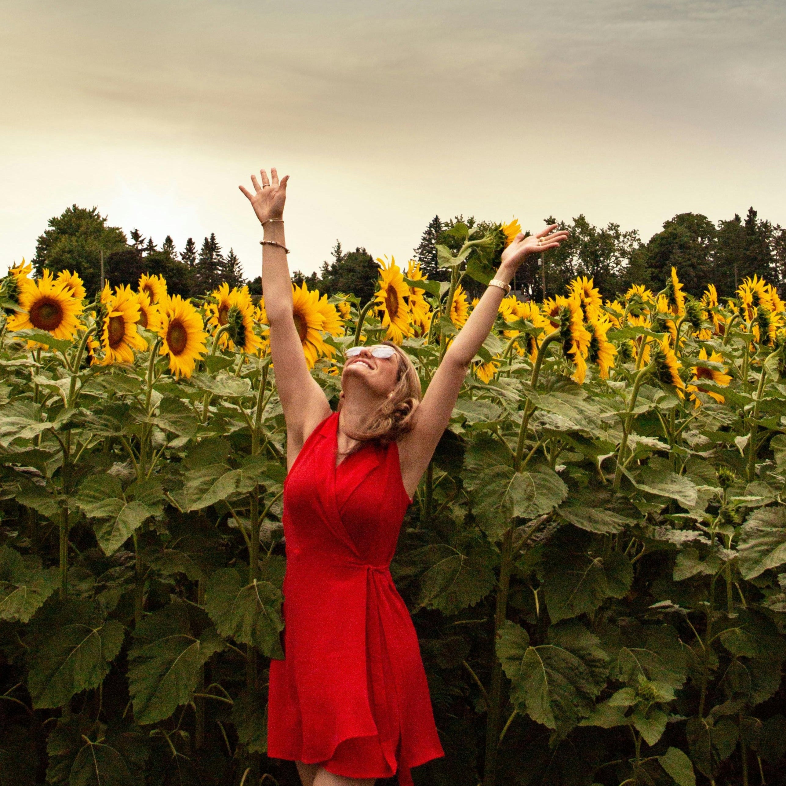 Woman in a red dress throwing a hat in a sunflower field with a blue sky.