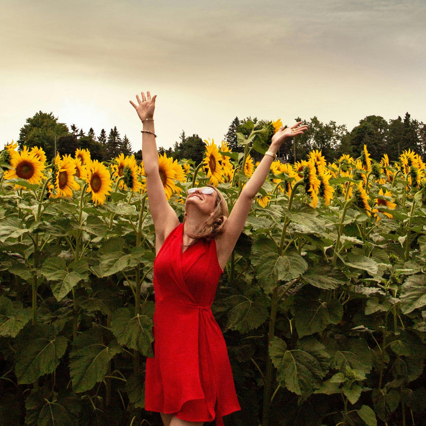 Woman in a red dress throwing a hat in a sunflower field with a blue sky.